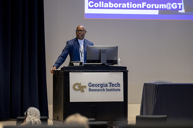 Presenter standing behind a podium with Georgia Tech Research Institute signage, delivering a talk in an auditorium with a large screen displaying event information.